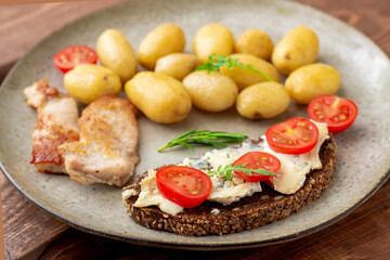 Boiled young potatoes, chunks of grilled turkey meat and cereal bread with blue cheese and tomatoes on a plate on a wooden background.