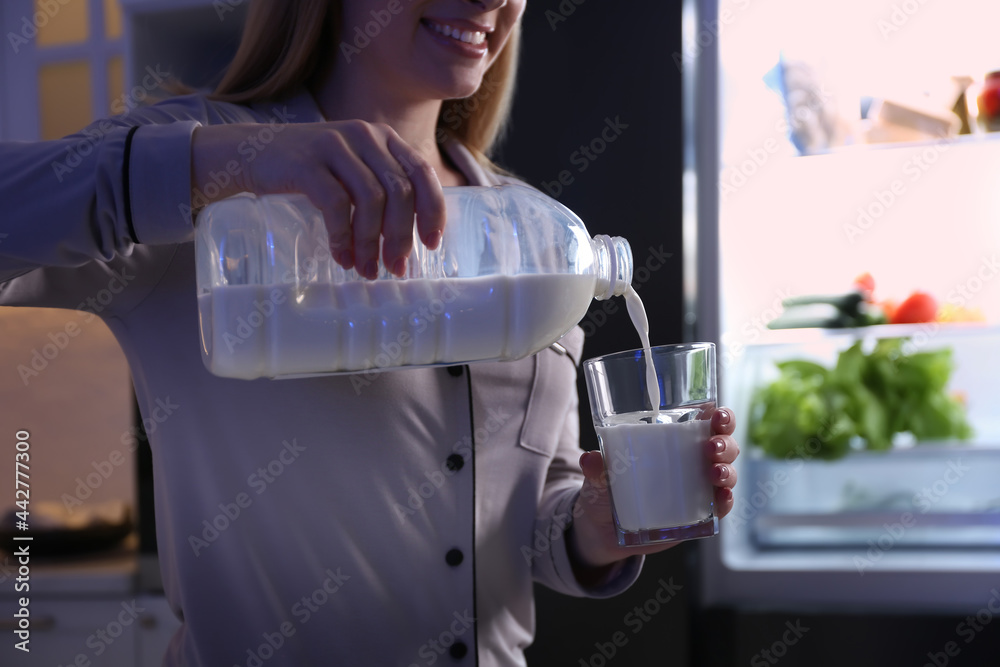 Sticker Young woman pouring milk from gallon bottle into glass near refrigerator in kitchen at night, closeup