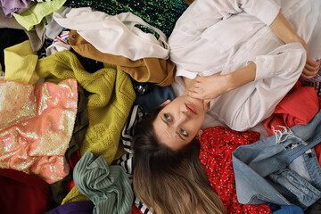 Young woman lying on pile of different clothes, top view. Fast fashion concept