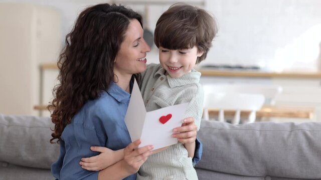 Happy young parent mom embrace cute kid son reading handmade postcard with red heart for mother day or valentines holiday. Millennial mommy hugging adorable preschool boy greeting with family event