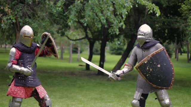 Battle of two medieval warriors in full sets of armor with swords and shields. Duel of knights in the park.