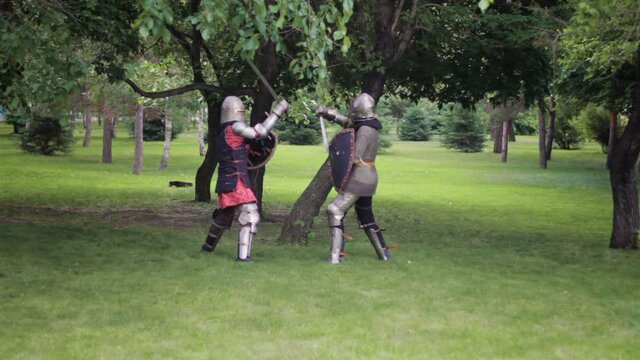 Battle of two medieval warriors in full sets of armor with swords and shields. Duel of knights in the park.