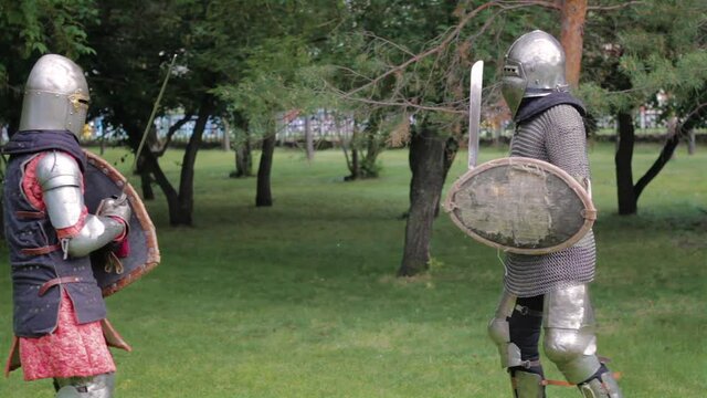 Battle of two medieval warriors in full sets of armor with swords and shields. Duel of knights in the park.