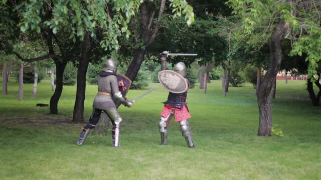 Battle of two medieval warriors in full sets of armor with swords and shields. Duel of knights in the park.