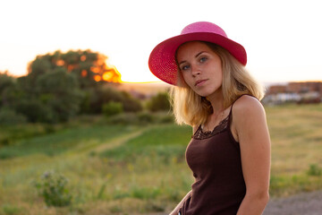 a girl in a pink hat and a brown T-shirt is standing in the sunset rays of the sun outside the city, looking at the camera