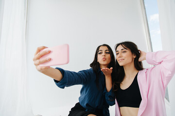 young armenian woman sending air kiss while taking selfie on smartphone with friend