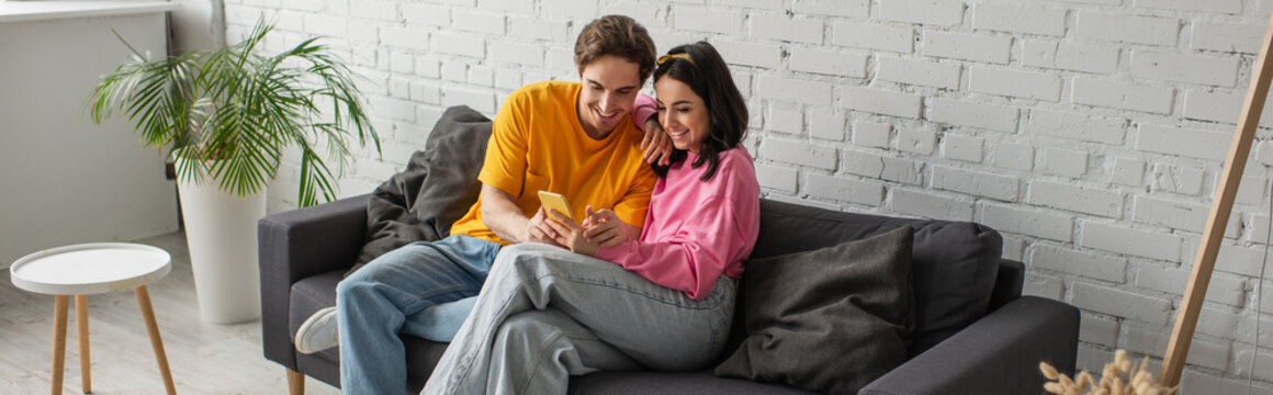 Smiling Young Couple Sitting On Couch And Looking At Cellphone In Living Room, Banner