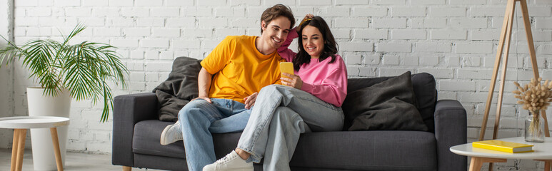 positive young couple sitting on couch and looking at cellphone in living room, banner
