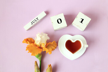 Calendar for July 1 : the name of the month of July in English, cubes with the number 0 and 1, a cup of tea in the shape of a heart, yellow iris on a pastel background, top view