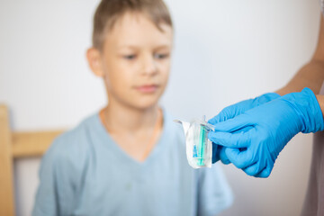 hands of a nurse in blue gloves open a pack of syringe close-up in front of the face of a boy in a blue t-shirt