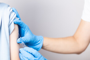Hands of a nurse in blue gloves injects a vaccine through a syringe into the shoulder of a brave boy in a blue T-shirt close-up on a white background