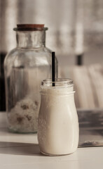 Milk shake, smoothie, in the glass mud on the white wooden table and glass bottle with white fluffy blow balls, dandelions, beige striped cushions in the background