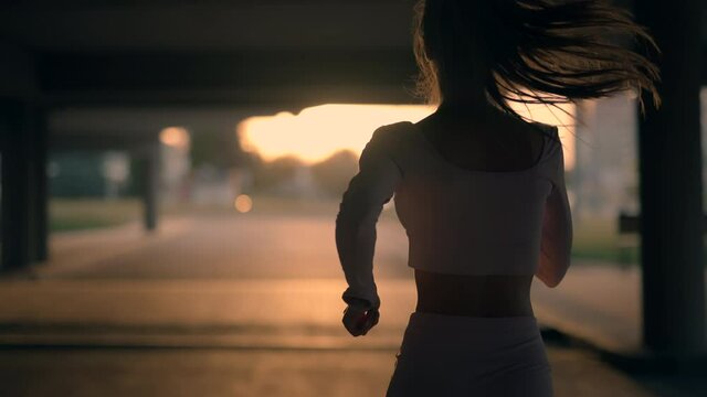 A young woman runner is training in the summer within a city. Close up of silhouette fitness girl is jogging outdoor. Concept of workouts running and healthy lifestyle. Back view. Slow motion.