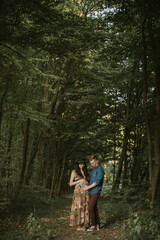 Fototapeta premium Happy Caucasian family, gorgeous pregnant young woman and cheerful handsome man, walking together in beautiful forest. Man and woman touching pregnant belly