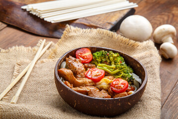 Udon with meat and vegetables in yakiniku sauce in a plate of coconut shells on a linen napkin next to chopsticks and vegetables.