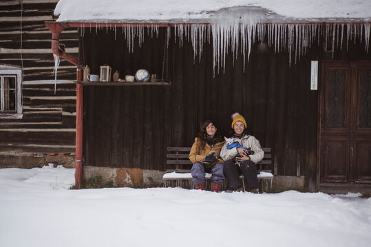 Queer female couple sit together in winter hut with icicles and dogs