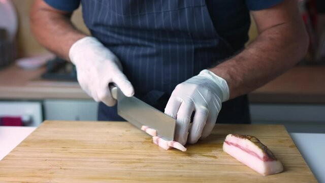 Male cook or chef cuts the bacon guanciale into thin strips on a wooden cutting board in a restaurant or at home.