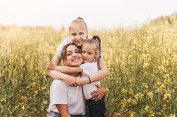 Naklejka na ściany i meble Happy young mother cuddles with her daughters in the summer on the field of rape. Happiness with family