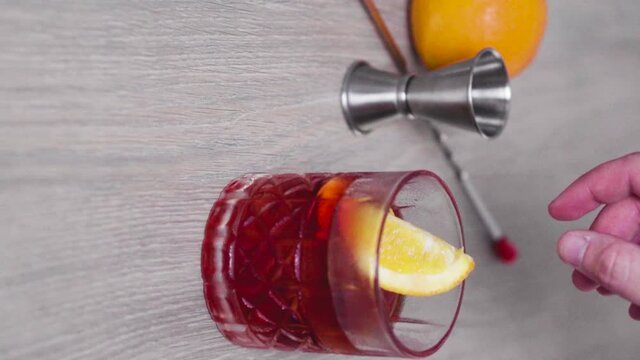 Slow motion of an ice-filled glass of negroni while dropping a piece of fresh orange in it.
