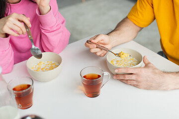 partial view of young couple sitting at table and eating corn flakes near cups of tea at home
