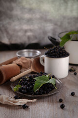 Black currant berries  in white cup and metal plate on wooden table in rustic traditional kitchen with wooden kitchen cutlery, vertical, selective focus, copy space
