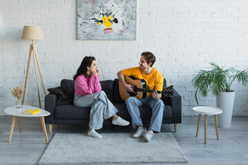 smiling young man sitting on couch and playing acoustic guitar near girlfriend with hands near face at home