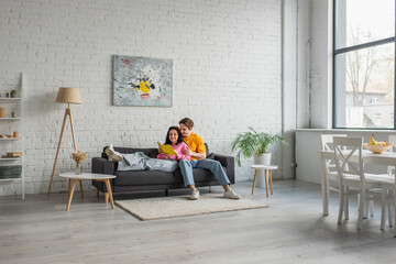 smiling young man hugging girlfriend lying on couch with book in hands in living room