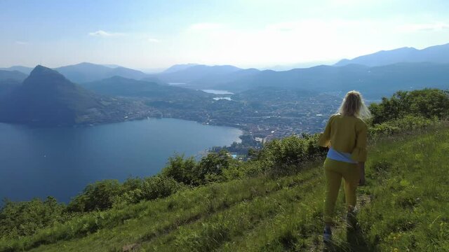 Swiss elegant woman walking on top of Lugano corniche by Lugano Lake in Switzerland. Aerial view lookout from Bre Mount. Lugano cityscape with San Salvatore mount in Ticino canton.