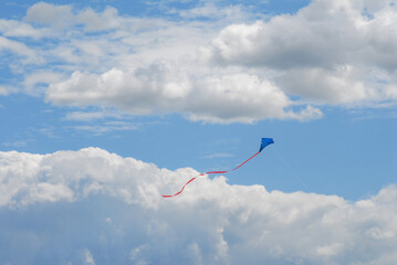 A blue kite with a long red tail flies high in the blue sky with beautiful white clouds. Blurred. A...
