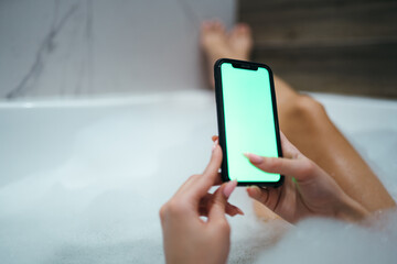 Photo of female legs lying in a foam bath and hands holding a phone with a light screen. Space for text