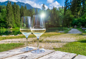 Two glasses of Prosecco white wine on a wood table with the background of a small alpine lake and the dolomite peaks of Trentino, Italy. Taken in late afternoon during summer 