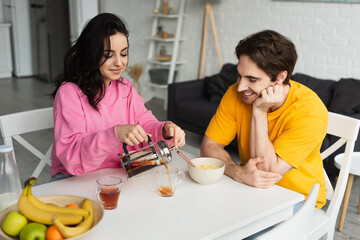 Obraz na płótnie Canvas smiling young woman sitting at table near boyfriend with breakfast and pouring tea from french press into cup in living room