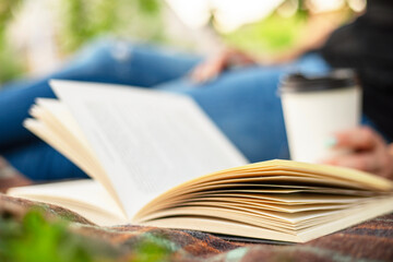 A woman in jeans with a book and a cup of coffee sits on a blanket in the park