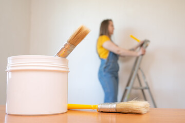 defocused woman on ladder looks dreamily in a room with an unpainted wall. Paint jar and brush on the foreground. selective focus.