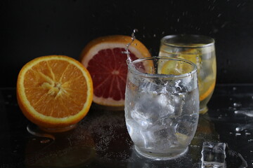 splashes of water over a glass of water and ice. Nearby are fruits orange and grapefruit, ice. On a black background. Refreshing cold ice drink