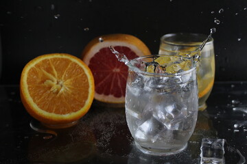 splashes of water over a glass of water and ice. Nearby are fruits orange and grapefruit, ice. On a black background. Refreshing cold ice drink