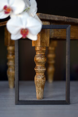 Close-up in an empty frame, the leg of an old carved wooden stool with an orchid branch descending from above. Vertical classics!