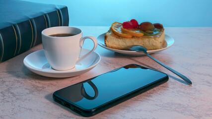 mobile phone, a cup of tea on a saucer and a piece of delicious homemade cake on a plate on a wooden kitchen table