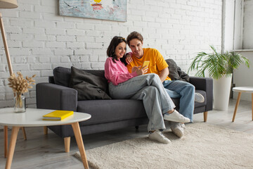 positive young couple sitting on couch and holding cellphone in living room - obrazy, fototapety, plakaty
