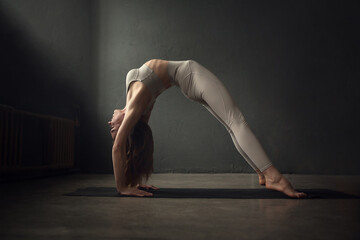 A slim beautifully lighted young  woman is doing Upward Bow (Wheel) yoga pose (Urdhva Dhanurasana) in a dark room. Image with selective focus and toning