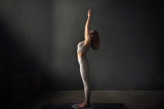 A slim beautifully lighted young  woman is doing Urdhva Hastasana yoga pose in a dark room. Image with selective focus and toning