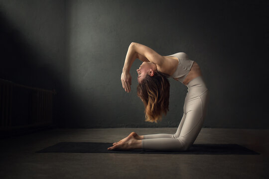 A Slim Beautifully Lighted Young  Woman Is Doing Camel Yoga Pose (Ustrasana) In A Dark Room. Image With Selective Focus And Toning