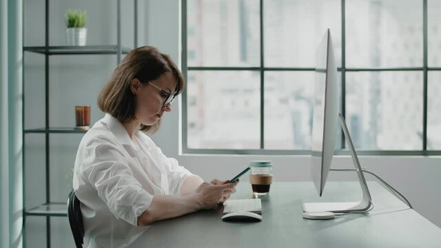 Businesswoman writes message on phone near computer monitor