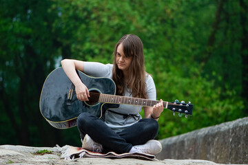 Pretty teenager girl playing guitar while sitting on the stone bridge. Young female guitarist outdoor in the nature