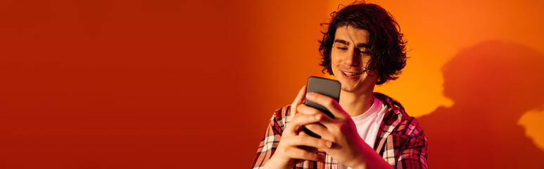 Cheerful curly man using mobile phone on orange background, banner