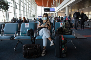 Coronavirus and social distancing. Traveling during pandemic. Young woman wearing a protective mask, waiting in the airport while looking at her smartphone. - obrazy, fototapety, plakaty