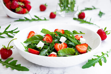 Summer salad of strawberry with arugula, spinach leaves, feta cheese in white plate on marble table with salad leaves and fresh strawberry fruits around. Strawberry salad recipe DIY. Step 3.