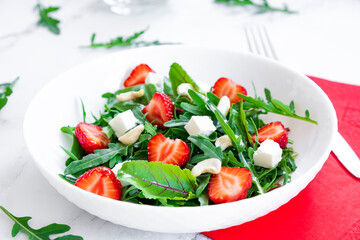 Salad of strawberry with arugula, spinach leaves, feta cheese, balsamic vinegar and cashew nuts with red napkin, salad leaves and fresh strawberry fruits around. Healthy food and raw food diet..