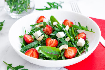 Vitamin salad of strawberry with arugula, young beet tops, feta cheese and cashew nuts in white plate with red napkin, salad leaves and fresh strawberry fruits around. Fruit detox and antioxidants.