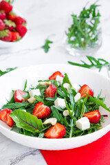Vitamin salad of strawberry with arugula, young beet tops, feta cheese and cashew nuts in white plate with red napkin, salad leaves and fresh strawberry fruits around. Keto diet and paleo diet.